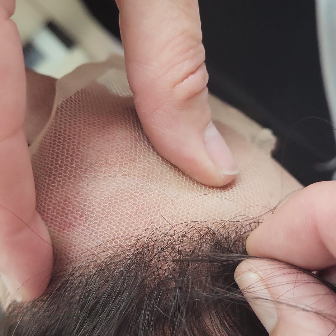 En esta imagen se muestra un primer plano de manos trabajando en la fabricación de una peluca. Se ve cómo se inserta cada hebra de cabello individualmente en una base de malla, creando una apariencia natural. Este proceso artesanal asegura que las prótesis capilares tengan rayas con efecto piel para una mayor naturalidad.